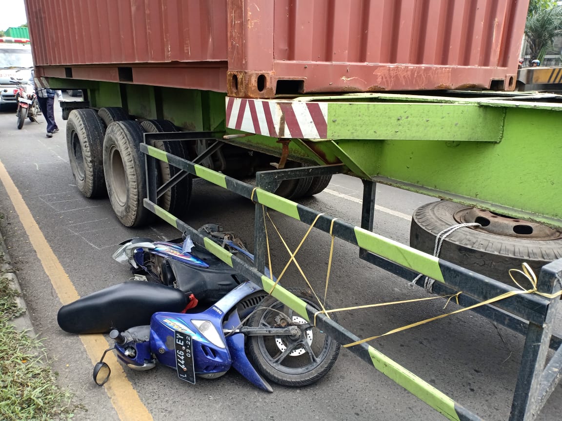 Posisi kendaraan berada di bawah truk trailer.